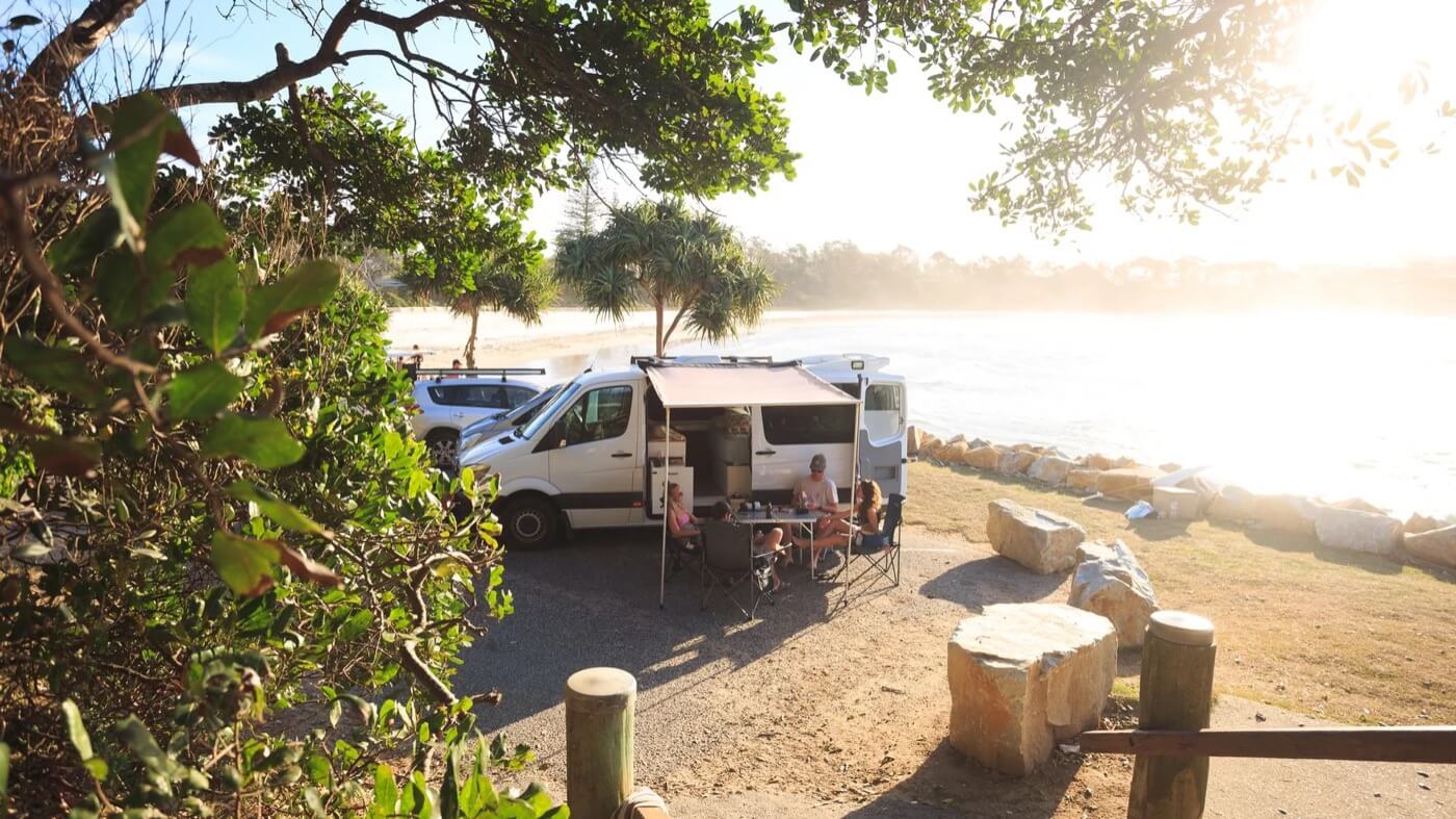 Van aménagé en bord de mer en Australie, avec une famille assise dehors sous l’auvent près de la plage